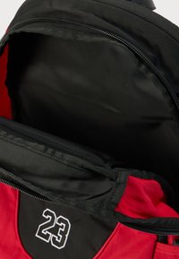 Κόκκινο και μαύρο σακίδιο πλάτης με ανοιχτό φερμουάρ που αποκαλύπτει το άδειο κύριο διαμέρισμα και ενισχυμένη θήκη για φορητό υπολογιστή, με τον αριθμό 23 σε λευκό χρώμα.