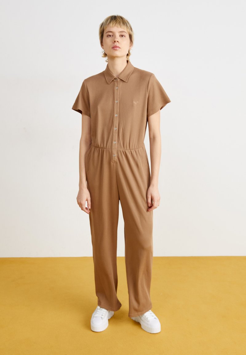 Combi-short marron à manches courtes, fermeture à boutons sur le devant, taille cintrée et jambes larges. Porté avec des baskets blanches à plateforme.