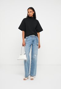 Frau trägt ein schwarzes, übergroßes Oberteil und helle, zerrissene Jeans, hält eine weiße Handtasche und steht vor einem weißen Hintergrund.