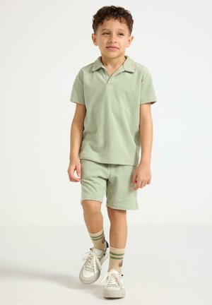 Nuori poika, jolla vaaleanvihreä poolopaita ja siihen sopivat shortsit, beige raidalliset sukat ja valkoiset tennarit, seisoo valkoisella taustalla.