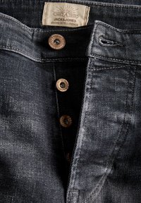 Close-up van een losgeknoopte donkergrijze spijkerbroek met zichtbare metalen knopen en een wit Jack & Jones-label op de tailleband.