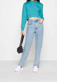 Teal kortärmad sweatshirt med rund halsringning och logopatch, matchad med ljusblå raka jeans. Modellen håller en liten svart väska.