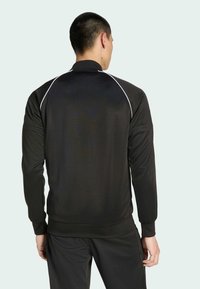 Μαύρο αθλητικό μπουφάν με ψηλό γιακά, μακριά μανίκια και λευκές λεπτομέρειες στους ώμους. Το υλικό φαίνεται λείο και ελαφρύ.