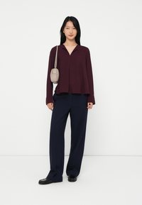 Woman standing wearing a burgundy long-sleeve blouse, navy wide-leg pants, black shoes, and carrying a small beige shoulder bag.