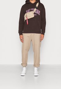 Felpa marrone con testo rosa, chinos beige e sneakers bianchi. Borsa a tracolla in tessuto beige con accenti rossi. Look casual streetwear.