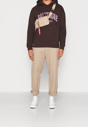 Bruine hoodie met roze tekst, beige chino's en witte sneakers. Schoudertas van beige stof met een rood accent. Casual streetwear look.