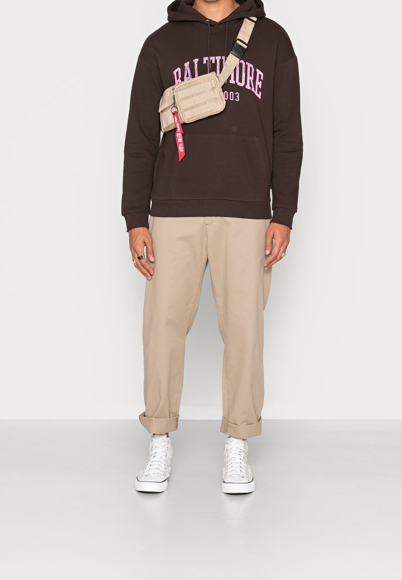 Felpa marrone con testo rosa, chinos beige e sneakers bianchi. Borsa a tracolla in tessuto beige con accenti rossi. Look casual streetwear.