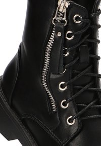 Bota de combate de cuero negro con cremallera lateral, parte delantera con cordones, ojales de metal y suela texturizada. Los detalles incluyen una costura visible.