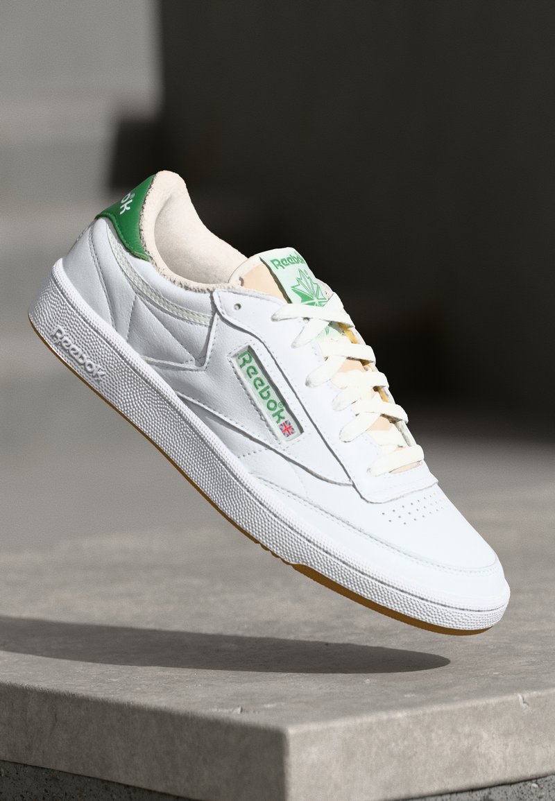 Sapatilha de couro branco com detalhes em verde, apresentando uma superfície texturizada, atacadores brancos e sola de borracha caramelo. Logotipo exibido na lateral.