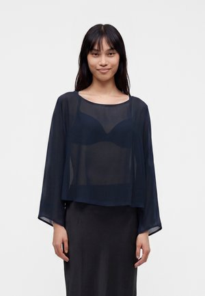 Femme aux longs cheveux foncés porte un haut bleu marine transparent à manches longues par-dessus un soutien-gorge noir, associé à une jupe noire, debout devant un fond uni.