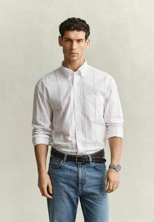 Junger Mann trägt ein weißes Hemd mit dünnen Streifen, blaue Jeans, einen braunen geflochtenen Gürtel und eine Armbanduhr, stehend vor einem schlichten Hintergrund.
