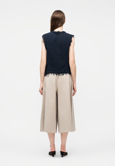 Γυναίκα που στέκεται με την πλάτη γυρισμένη, φορώντας αμάνικο μπλε σκούρο υφασμάτινο τοπ με κρόσσια στις άκρες και μπεζ φαρδύ παντελόνι cropped με μαύρες ψηλοτάκουνες γόβες.