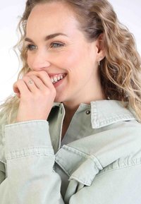 Eine hellgrüne Jeansjacke mit Knopfverschluss und umgeschlagenen Ärmeln. Der Träger trägt einen silbernen Ring und Ohrringe, lächelt und hat lockiges Haar.