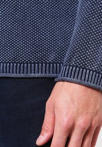 Hand nahe der Hüfte ruhend, trägt einen dunkelblauen strukturierten Strickpullover mit gerippten Bündchen und dunklen Hosen.