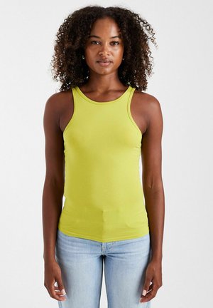 Femme aux cheveux bouclés portant un haut sans manches vert citron et un jean bleu clair, debout sur un fond blanc uni.