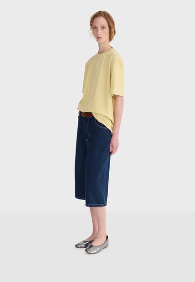 Jeune personne debout portant une chemise ample jaune à manches courtes, un pantalon large bleu foncé court avec une ceinture marron, et des ballerines argentées métalliques.