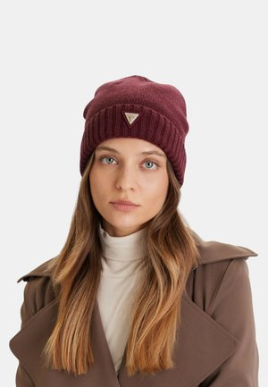 Donna con capelli lunghi castani che indossa un berretto di lana color bordeaux con un logo metallico triangolare, un dolcevita beige e un cappotto marrone.