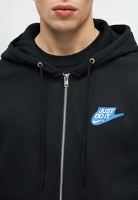 Άνδρας που φοράει μαύρο φούτερ με φερμουάρ και κορδόνια κουκούλας, με μπλε λογότυπο Nike "JUST DO IT." στην αριστερή πλευρά του στήθους.