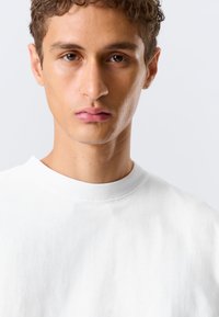 Jeune homme aux cheveux bouclés et portant une boucle d'oreille, vêtu d'un t-shirt blanc uni à col rond, regardant directement l'appareil photo sur un fond clair.