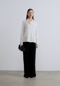 White button-up shirt with long sleeves, paired with a black maxi skirt. Accessorized with a small black handbag. Smooth fabric texture.
