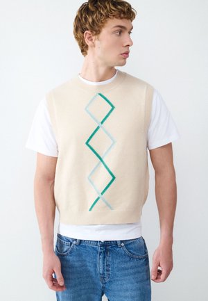 Junger Mann trägt einen beigen ärmellosen Pullover mit grünen und blauen Rautenmuster über einem weißen T-Shirt und blauen Jeans, schaut zur Seite.