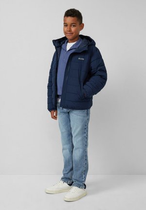 Blauwe pufferjack met capuchon, rits aan de voorkant en reliëf SLVR-logo; gecombineerd met lichtblauwe jeans en witte sneakers.