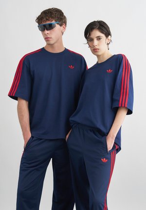 Dva modela oblečena v usklajene temno modre trenirke Adidas z rdečimi črtami in logotipi, ki stojita skupaj ob strani na enobarvnem ozadju.