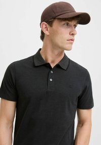 Zwarte polo met een gestructureerde kraag, een placket met twee knopen en een klein logo. Het model draagt een bruine baseballcap.