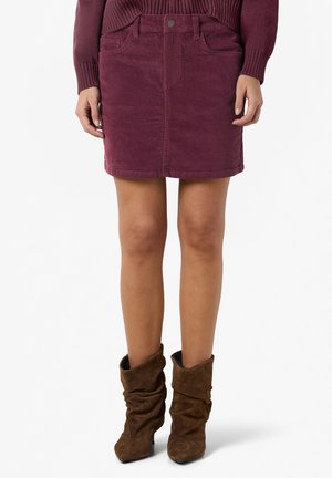 Femme portant une mini-jupe en velours côtelé bordeaux, un pull assorti et des bottines en daim marron décontractées, debout sur fond blanc.