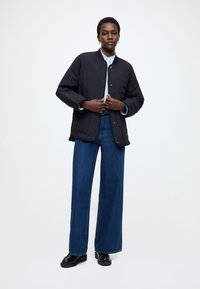 Crna prošivena jakna s prednjim zatvaračem na drukere, nošena preko svijetloplave košulje; uparena s visokim strukom, širokim nogavicama plavim trapericama i crnim cipelama.