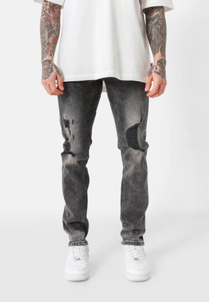Jean noir en denim usé avec des zones décolorées, des patchs visibles et une coupe fuselée, associés à des baskets blanches sur un fond uni.