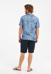 Kortebloesem met knopen in lichtblauw met een bloemenpatroon. Draagt een donkerblauwe korte broek en slippers, wat een ontspannen pasvorm laat zien.