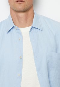 Lichtblauw overhemd met korte mouwen, gemaakt van textuurstof, met een knoopsluiting aan de voorkant en een enkele borstzak. Gedragen over een wit T-shirt met ronde hals.