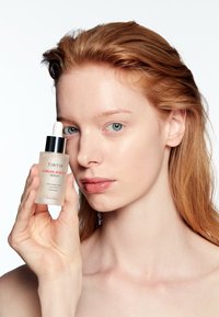 Giovane donna con pelle chiara tiene una bottiglia di siero all'acido azelaico TIRTIR 12% vicino al viso su uno sfondo bianco.