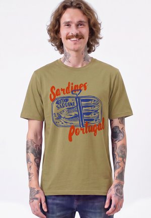 Young man with shoulder-length hair and tattoos wears olive green t-shirt with sardine can graphic and red text "Sardines Portugal."