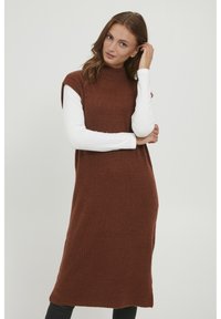Robe en tricot marron avec une texture côtelée et un col montant. Superposée à un haut à manches longues blanc, elle présente une silhouette décontractée et ample.