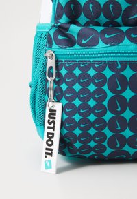 Σακίδιο της Nike με μοτίβο σε τιρκουάζ και ναυτικό μπλε, διακοσμημένο με κυκλικούς λογότυπους. Περιλαμβάνει ασημένιο φερμουάρ και μια ετικέτα που γράφει "ΑΠΛΑ ΚΑΝΕ ΤΟ".