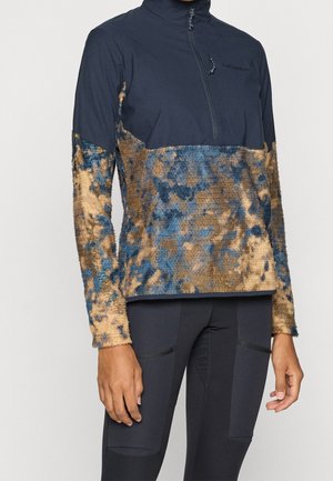 Persoon draagt een marineblauwe en bruin gevlekte fleece trui met een korte ritssluiting en zwarte legging met gestikte zakdetails.