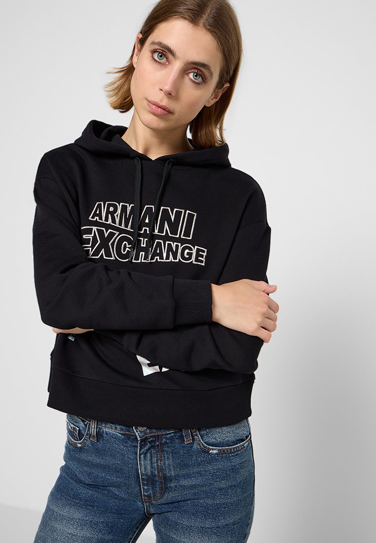 Sudadera negra con capucha, con puños y dobladillo acanalados, que presenta un logo blanco prominente en la parte frontal. Combinada con jeans de mezclilla azul.