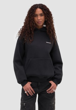 Vrouw met krullend haar die een zwarte WRSTBHVR-hoodie en zwarte cargobroek draagt, staand met handen vlak bij haar zakken tegen een effen achtergrond.