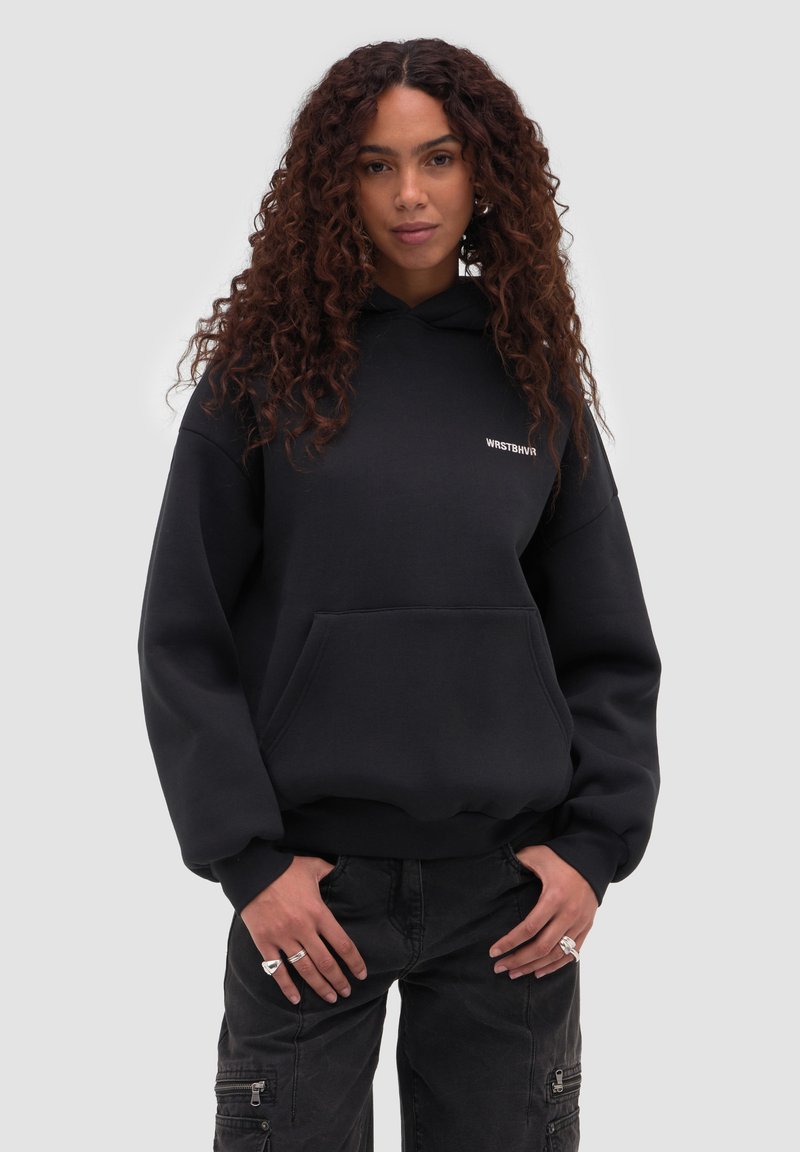 Donna con capelli ricci che indossa una felpa nera WRSTBHVR con cappuccio e pantaloni cargo neri, in piedi con le mani vicino alle tasche davanti a uno sfondo semplice.