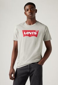 Siva pamučna majica s crvenim Levi's logom u zakrivljenom dizajnu, uparen s crnim trapericama. Klasični okrugli izrez i kratki rukavi.