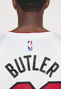 Balta basketbola krekls ar melnu uzrakstu "BUTLER" un sarkanām detaļām. Aizmugurē ir NBA logo birka, trīstūra formas apkakle un strukturēta auduma faktūra.