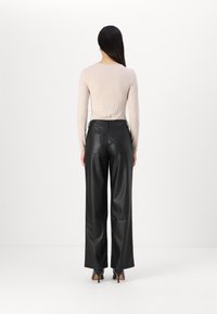 Zwarte faux leren wijde broek met twee achterzakken, gecombineerd met een lichtbeige top met lange mouwen. Model staat met de rug naar de camera.