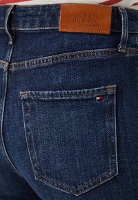 Jeans de denim azul oscuro con costuras marrones, bolsillo trasero con pequeño logotipo de la bandera de Tommy Hilfiger y parche de cuero de la marca Tommy Hilfiger en la cinturilla.