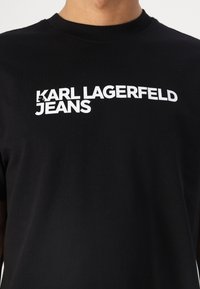 Juodas medvilninis marškinėliai su ryškiu baltu tekstu "KARL LAGERFELD JEANS" ant krūtinės. Apvalus kaklas, trumpomis rankovėmis.