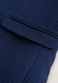 Primer plano de tela azul oscuro con un pequeño patrón a cuadros y un detalle de solapa doblada, probablemente parte de un bolsillo de prenda.