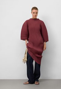 Burgunderfarbendes, strukturiertes Kleid mit übergroßen Ärmeln, asymmetrischem Saum, kombiniert mit dunklen Jeans und einer cremefarbenen Handtasche. Einfache Sandalen vervollständigen den Look.