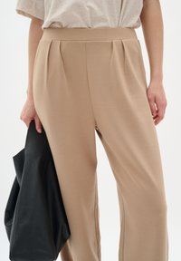 Beige, högmidjade byxor med veck vid midjan. Materialet verkar mjukt och slätt. En svart totebag hålls i handen.