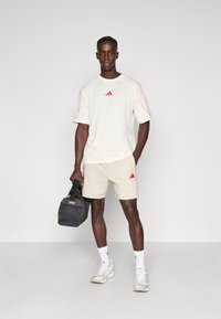 Σετ ανδρικής αθλητικής ένδυσης που περιλαμβάνει εκρού κοντομάνικο πουκάμισο με κόκκινες λεπτομέρειες και μπεζ σορτς, καθώς και μαύρη τσάντα duffle.